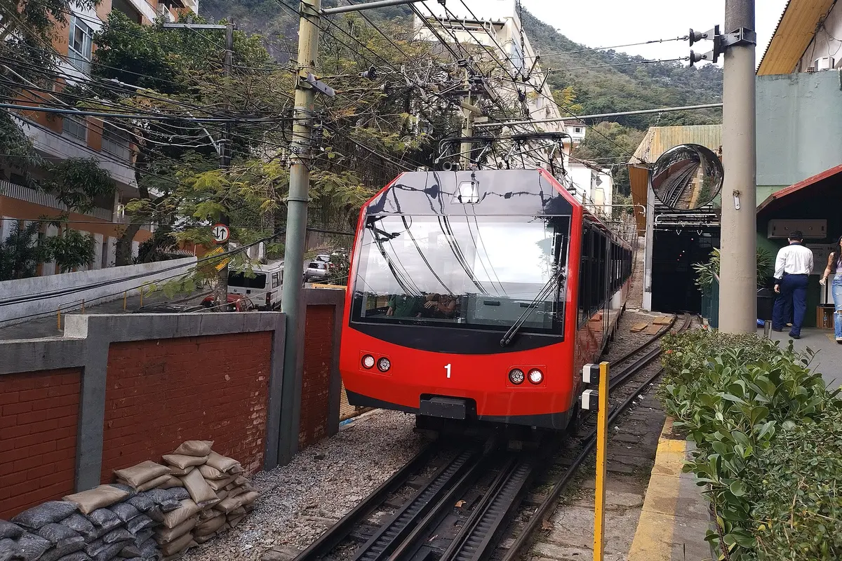 A red Corcovado cog train at Cosme Velho station, with the rack rail between the tracks and forested hills behind